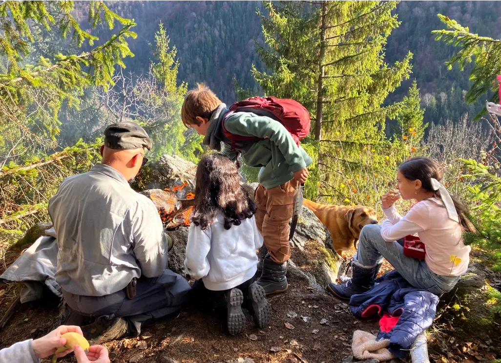 Children and an instructor gathered around a small campfire on a forested mountainside during a forest school session